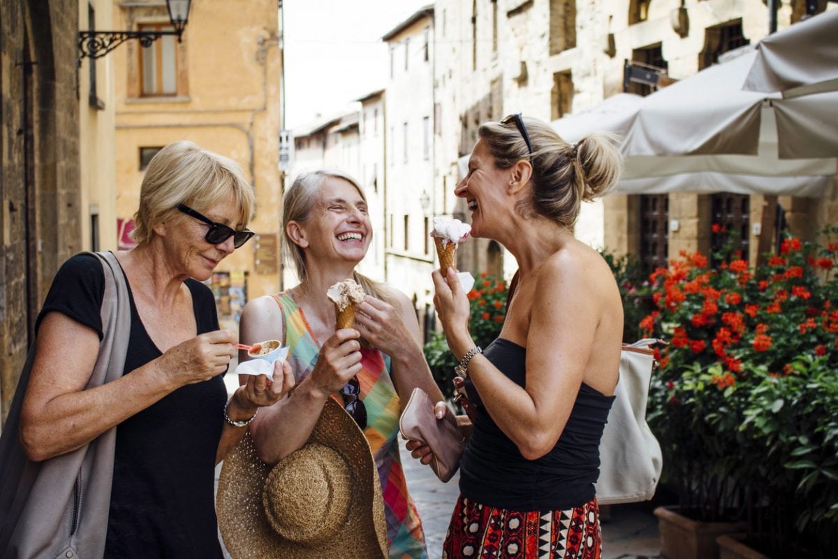 Three mature female friends standing eating Italian ice-creams while in a street in Tuscany during summer. They are smiling and facing each other and enjoying their holiday.